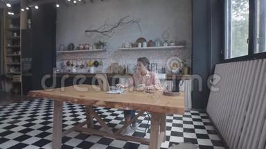 美丽的年轻女子坐在厨房的木桌旁，喝茶或咖啡，在她的银色笔记本电脑上打字